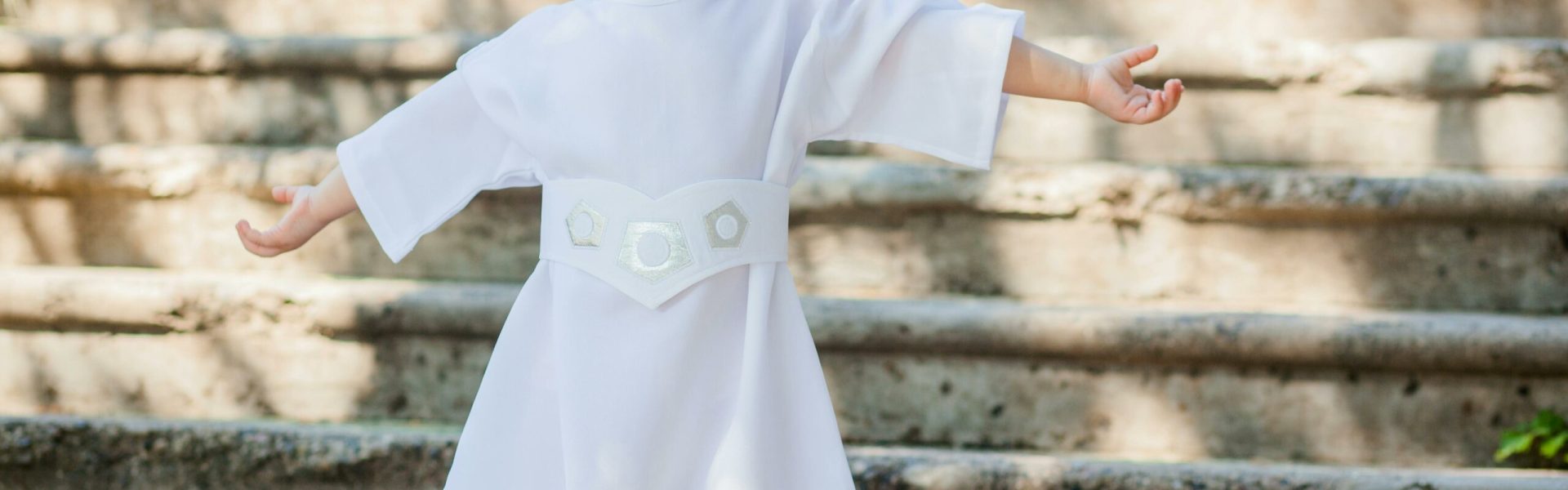 A little girl gleefully posing in a Princess Leia costume on outdoor steps during daytime.